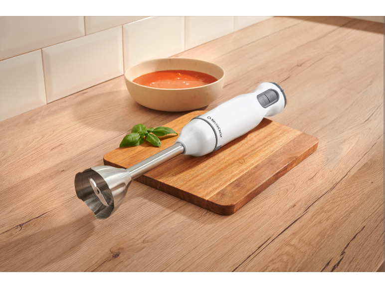 Rankinis trintuvas ant medinės lentos šalia pomidorų sriubos dubenėlio ir baziliko.