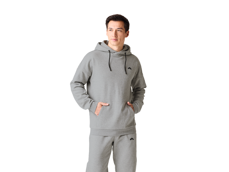 Vyras su pilku džemperiu su gobtuvu ir sportinėmis kelnėmis, rankos kišenėse.