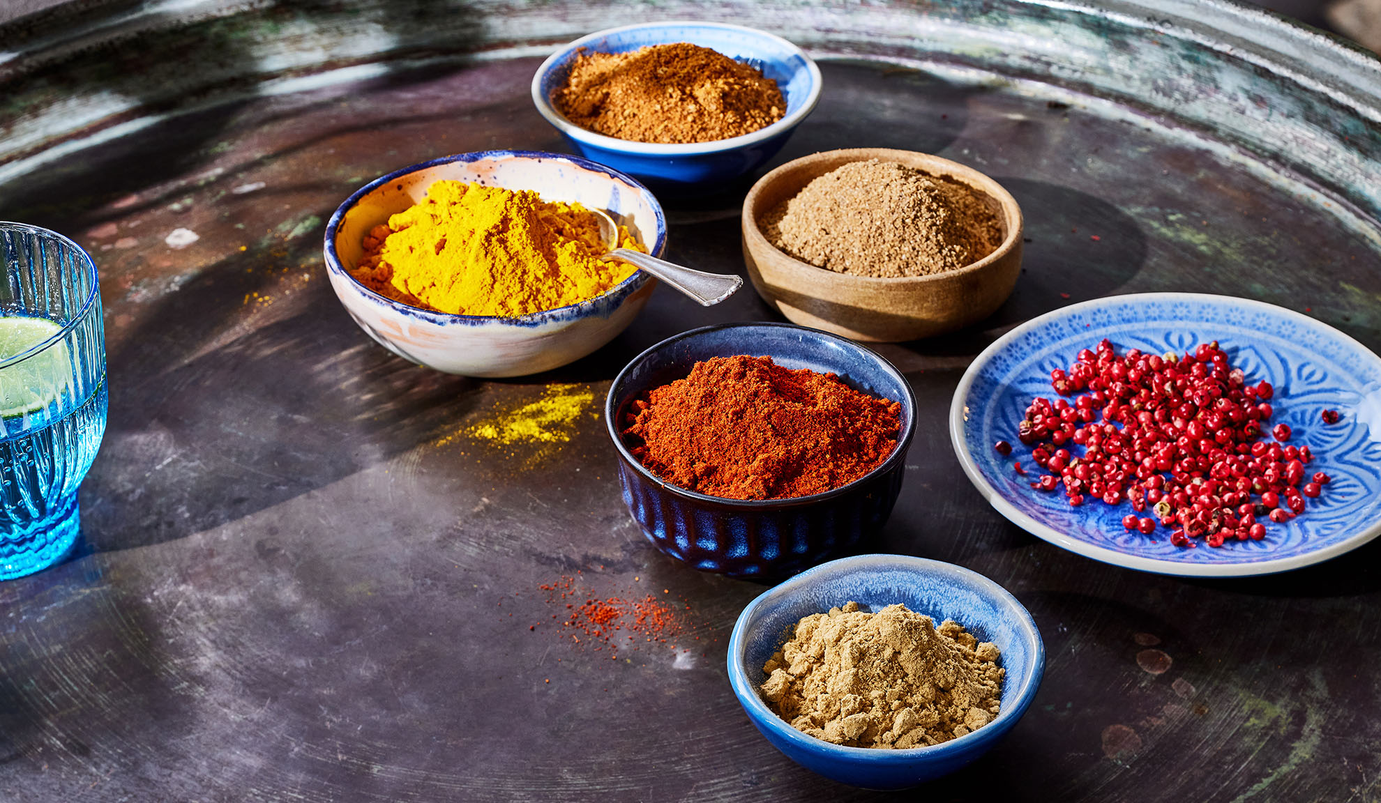 Įvairūs prieskoniai dubenėliuose: ciberžolė, paprika, pipirai ir kiti ant metalinio padėklo.