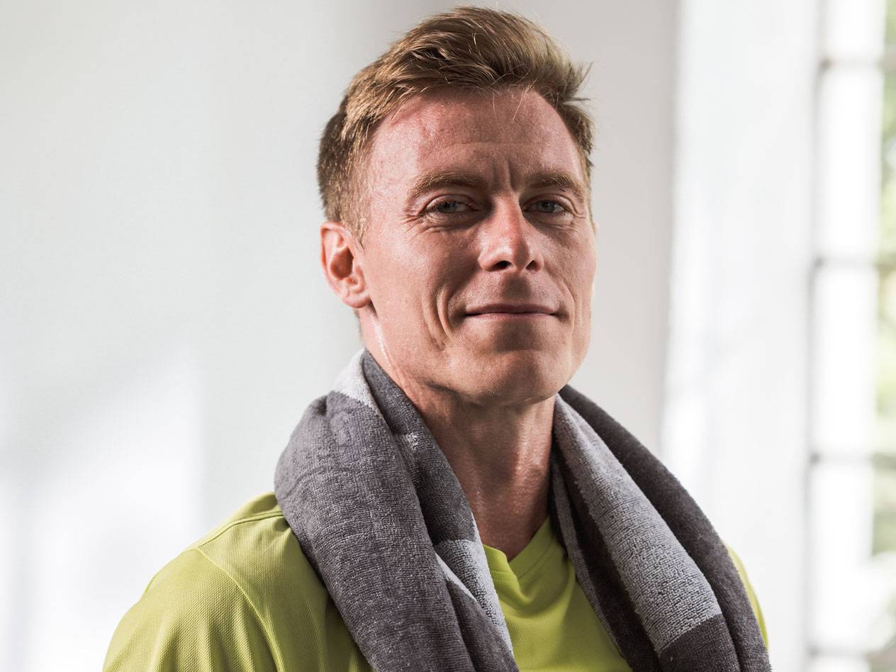 Šviesiaplaukis vyras su rankšluosčiu ant kaklo žiūri į kamerą.