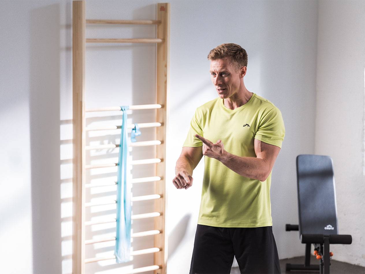 Vyras sportine apranga stovi prieš švedišką sienelę su pasipriešinimo juosta.