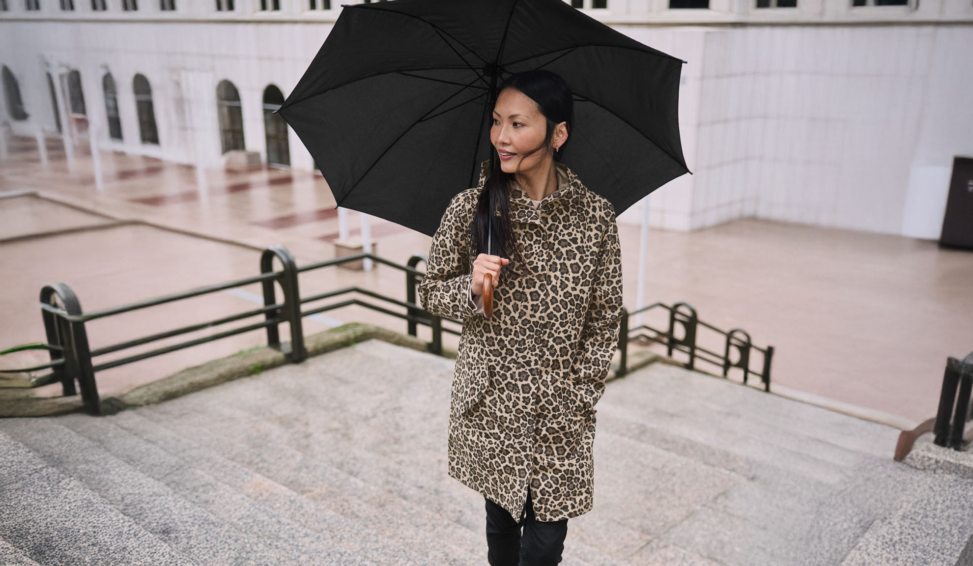 Moteris su leopardo rašto paltu laiko juodą skėtį ant laiptų.