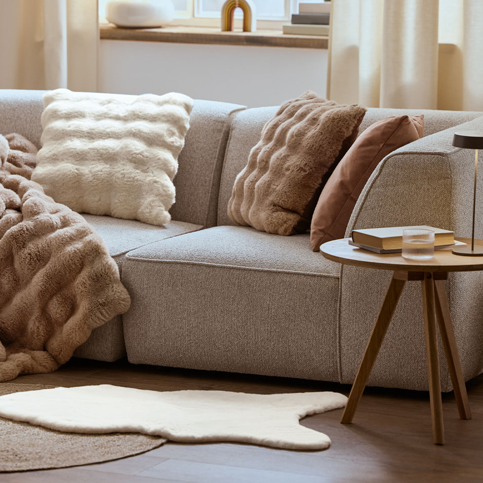 Pilka modulinė sofa su pūkuotomis pagalvėmis ir antklode, šalia medinio šoninio staliuko.