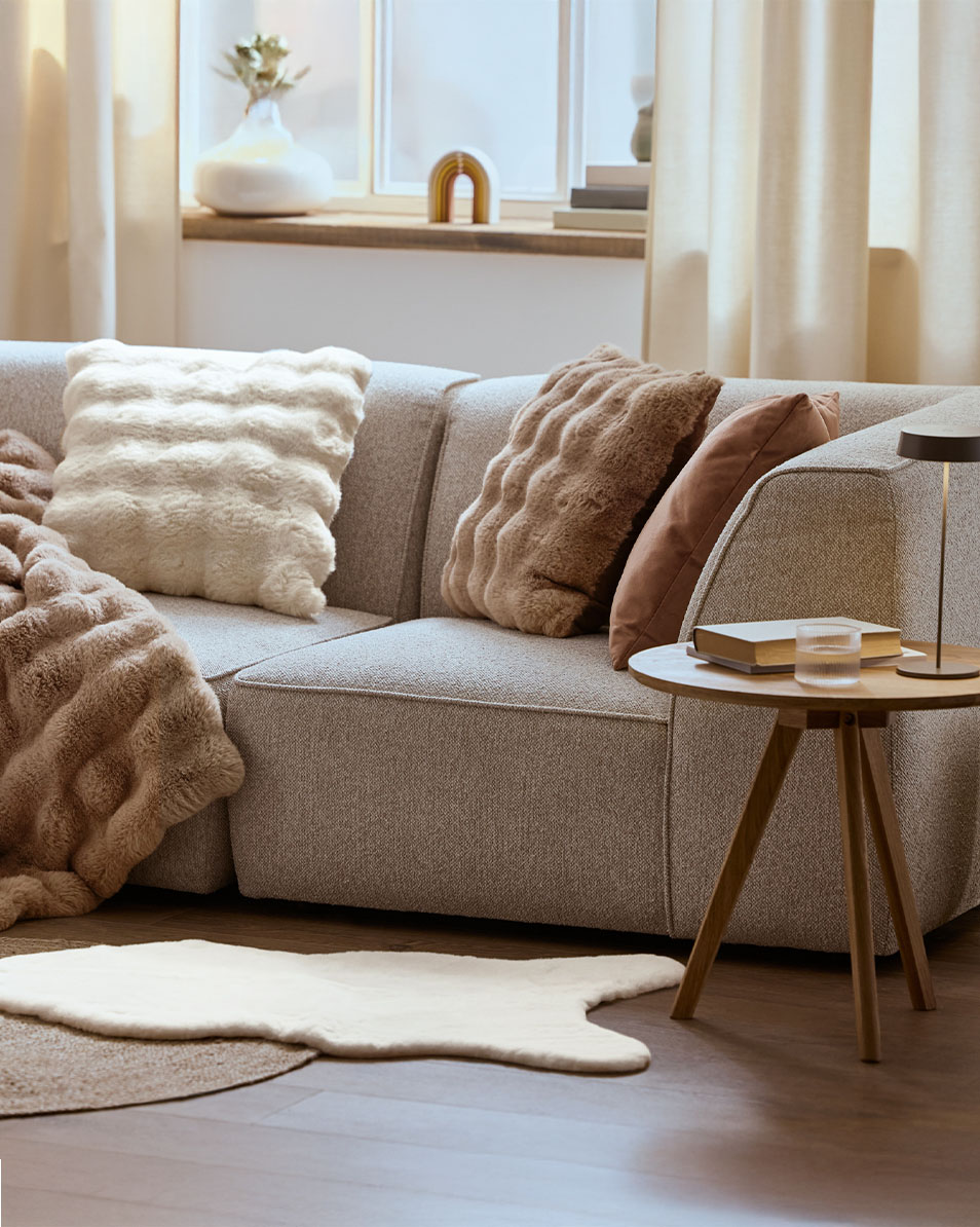 Pilka modulinė sofa su pūkuotomis pagalvėmis ir antklodėmis, medinis šoninis staliukas.