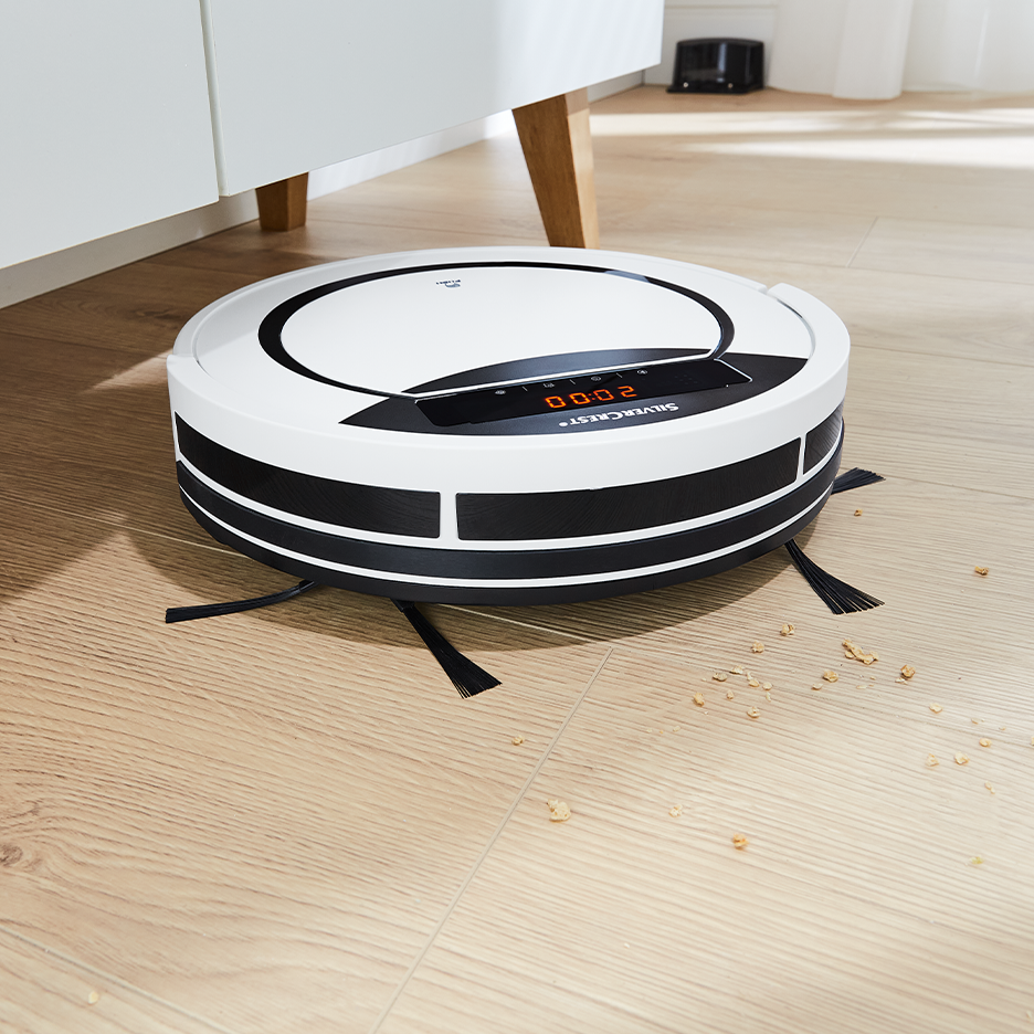 Baltas robotas dulkių siurblys ant medinių grindų su trupiniais.