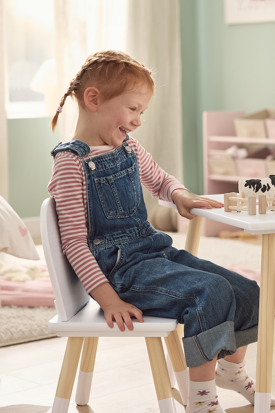 Linksma mergaitė sėdi prie balto vaikiško staliuko su kėdute, žaidžia su karvutės figūrėlėmis.