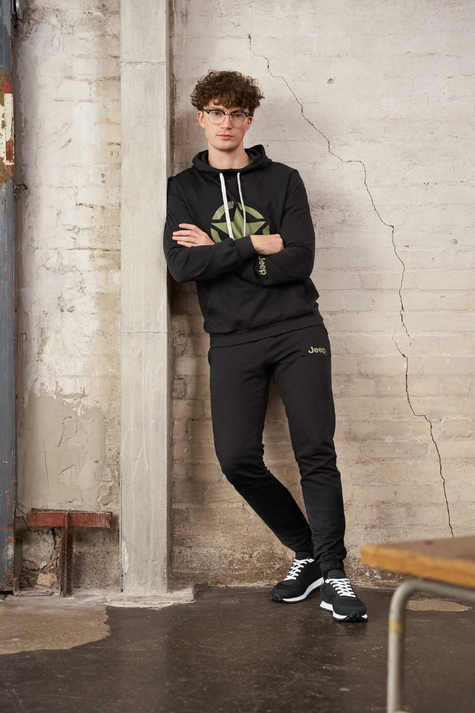 Vyras su juodu džemperiu su gobtuvu ir sportinėmis kelnėmis, atsirėmęs į sieną.