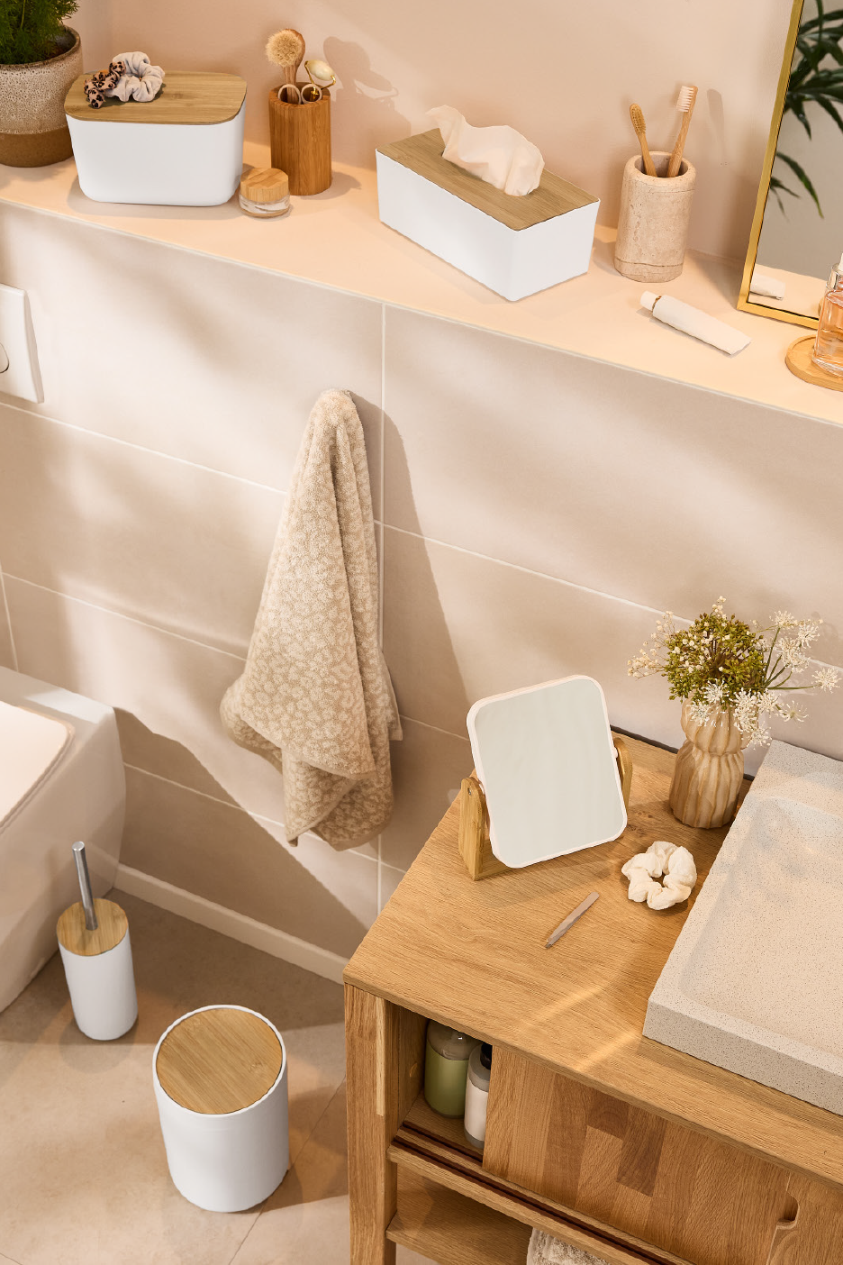 Vonios kambario aksesuarai: baltos dėžutės su bambukiniais dangteliais, rankšluostis, veidrodis ir medinė spintelė.