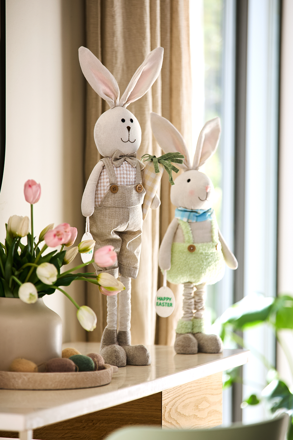 Du velykiniai zuikiai, tulpės ir dekoratyviniai kiaušiniai ant stalo. Ant zuikių kabo užrašai „Happy Easter“.