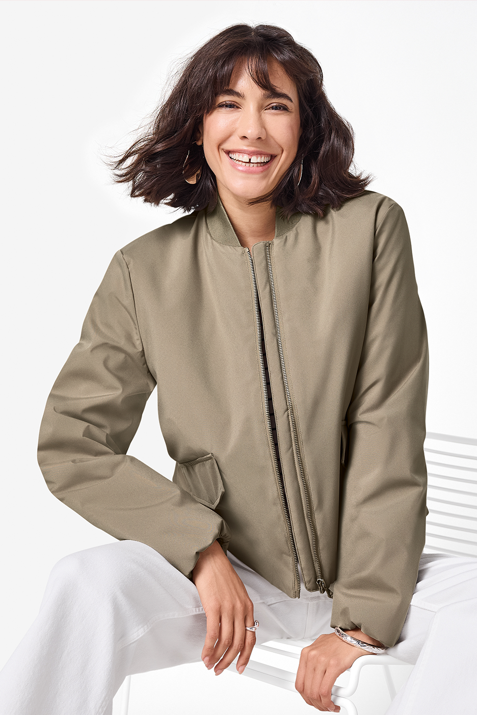 Moteris su chaki spalvos bombonešio striuke ir baltais džinsais šypsosi į kamerą.