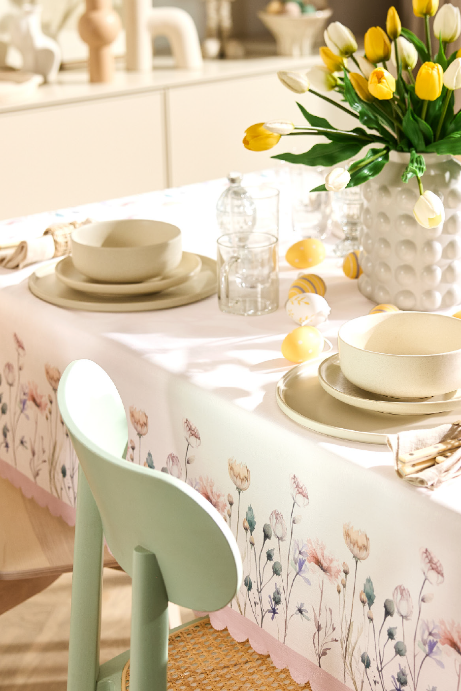 Velykinis stalas su gėlių staltiese, geltonomis tulpėmis ir indais.