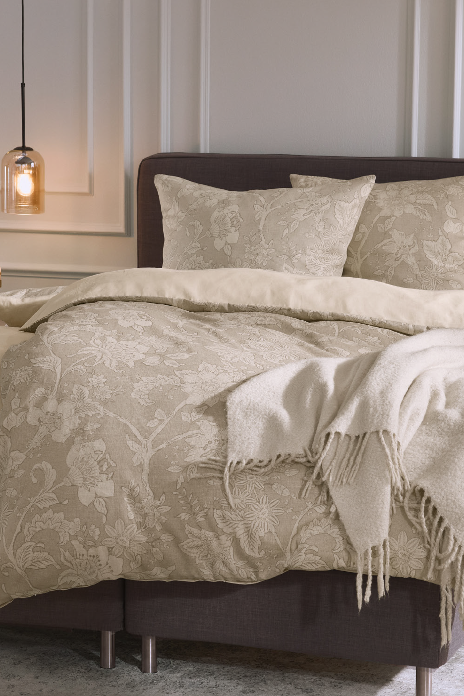 Šviesiai smėlio spalvos patalynės komplektas su gėlių raštais ant lovos su tamsiu galvūgaliu.