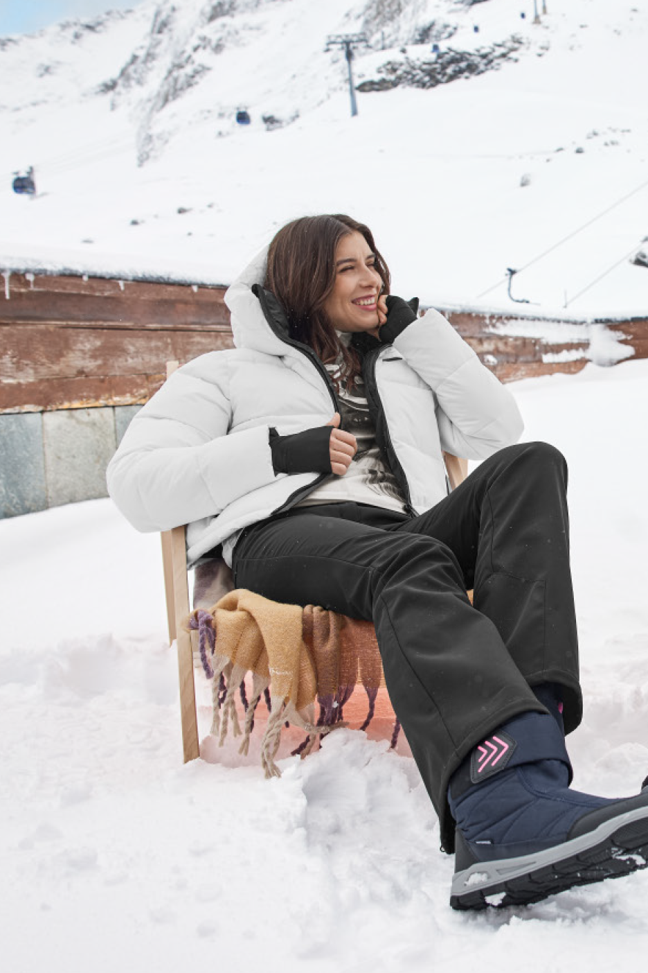 Moteris sėdi snieguotoje kalnų vietovėje, vilkėdama baltą pūkinę striukę ir juodas kelnes.