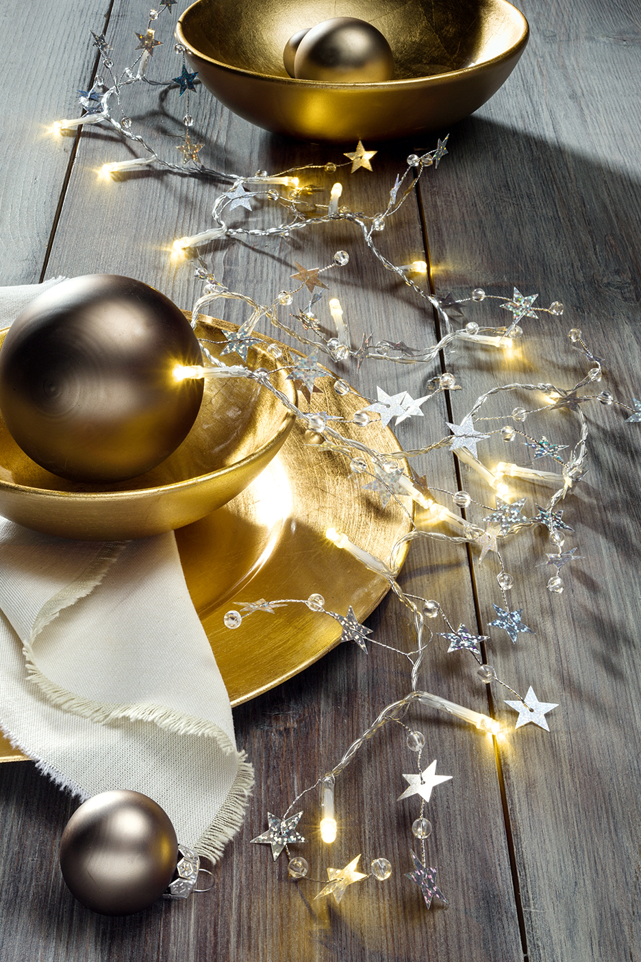 Šventinė dekoracija su auksiniais dubenėliais, matiniais rutuliais ir žvaigždžių girlianda su lemputėmis.