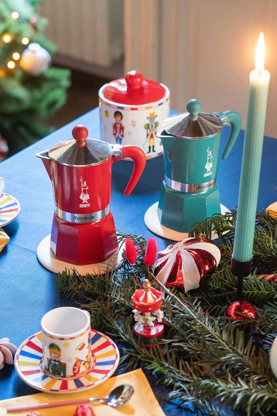Raudonas ir žalsvai mėlynas „Bialetti Rainbow“ kavos virimo aparatas ant stalo su kalėdinėmis dekoracijomis.