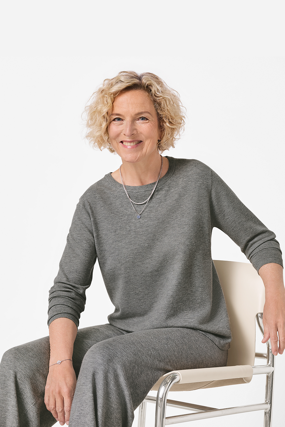 Moteris su pilku megztiniu ir kelnėmis sėdi ant kėdės ir šypsosi