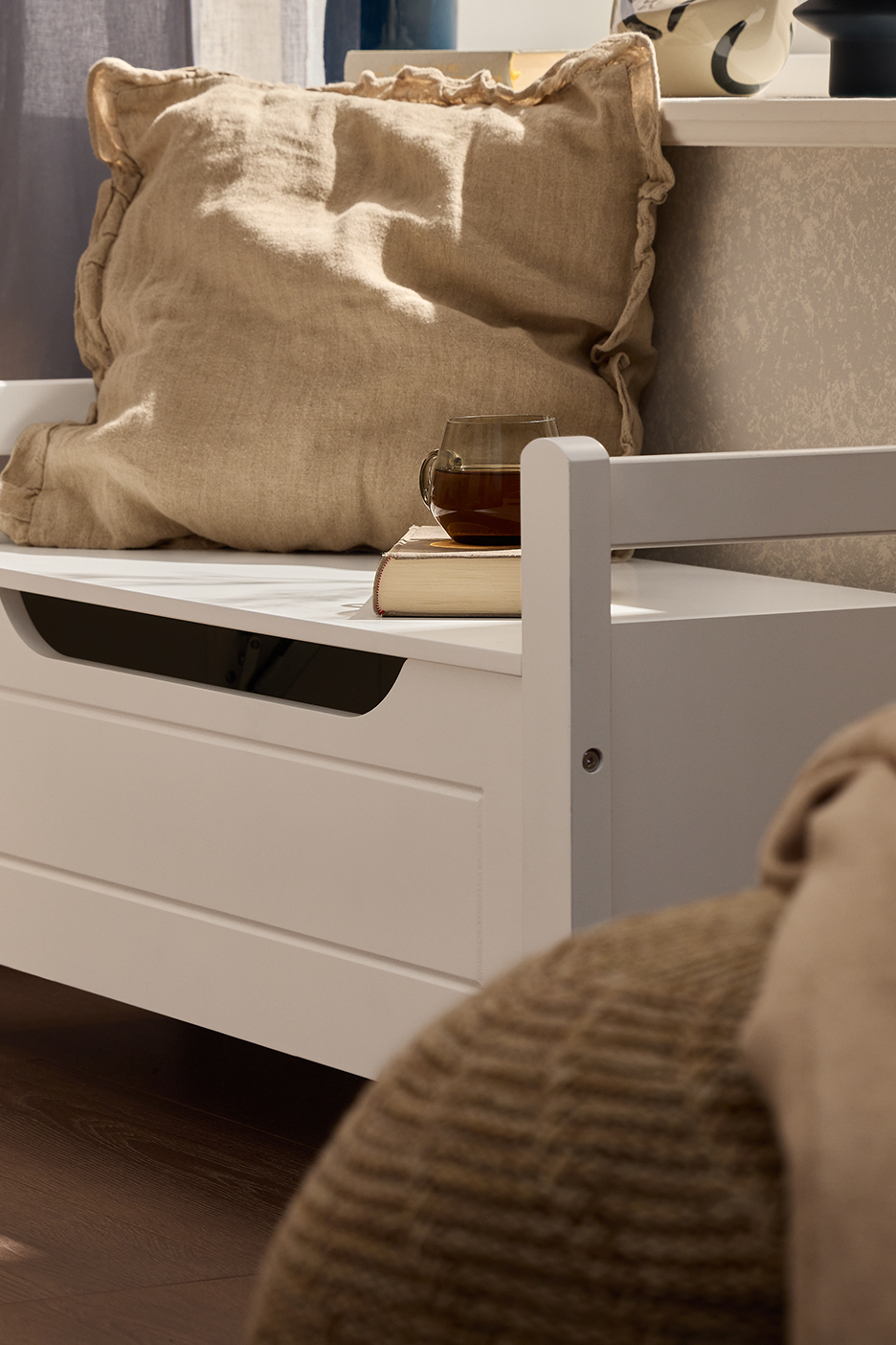 Balta daiktadėžė-suoliukas su smėlio spalvos pagalve, knyga ir puodeliu arbatos.