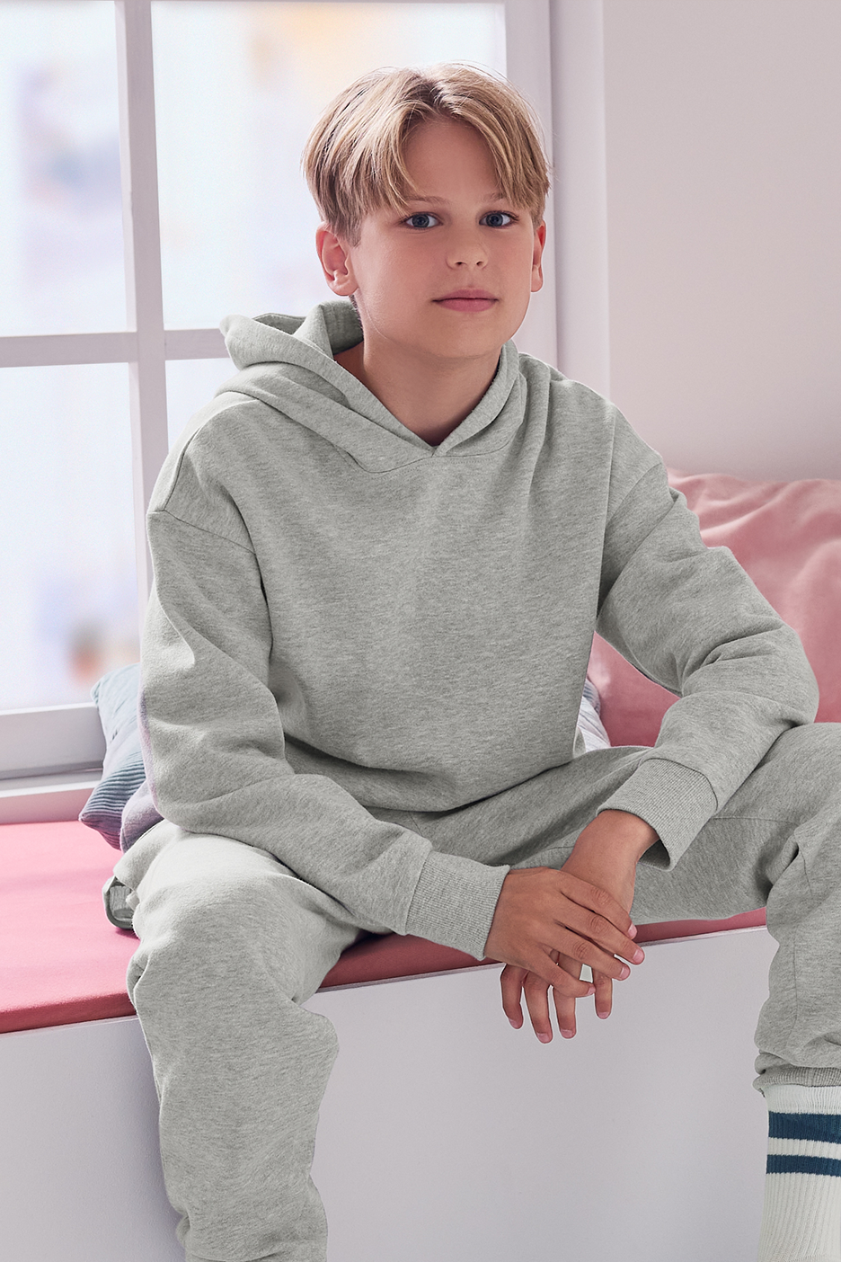 Berniukas, sėdintis ant palangės, vilkintis pilką džemperį su gobtuvu ir sportines kelnes.