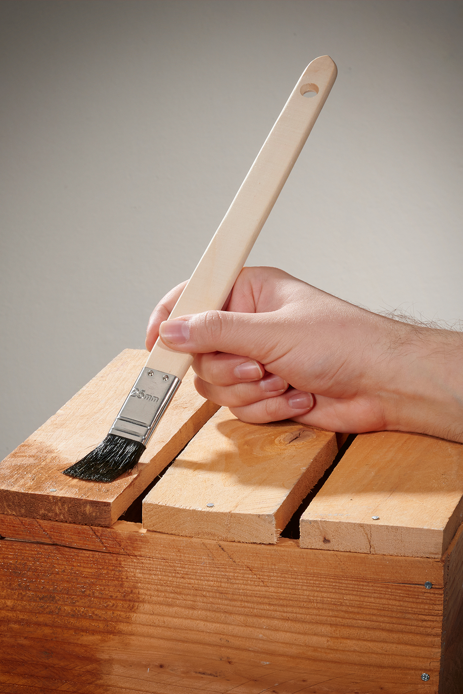 Rankos dažo medinę dėžę teptuku, ant kurio parašyta „25mm“.