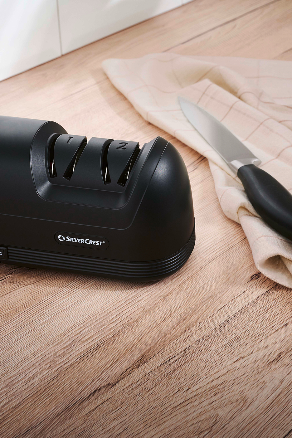 Juodas elektrinis peilių galąstuvas „SilverCrest“ su peiliu ir audiniu ant medinio paviršiaus.