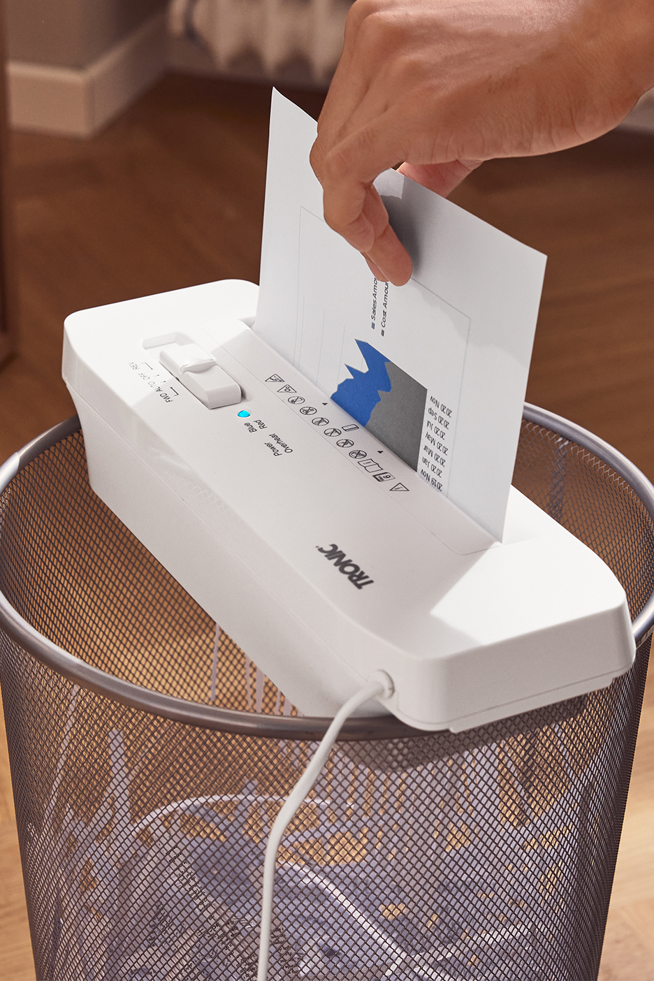 Asmuo naudoja baltą TRONIC popieriaus smulkintuvą, įdėtą į metalinę šiukšliadėžę.