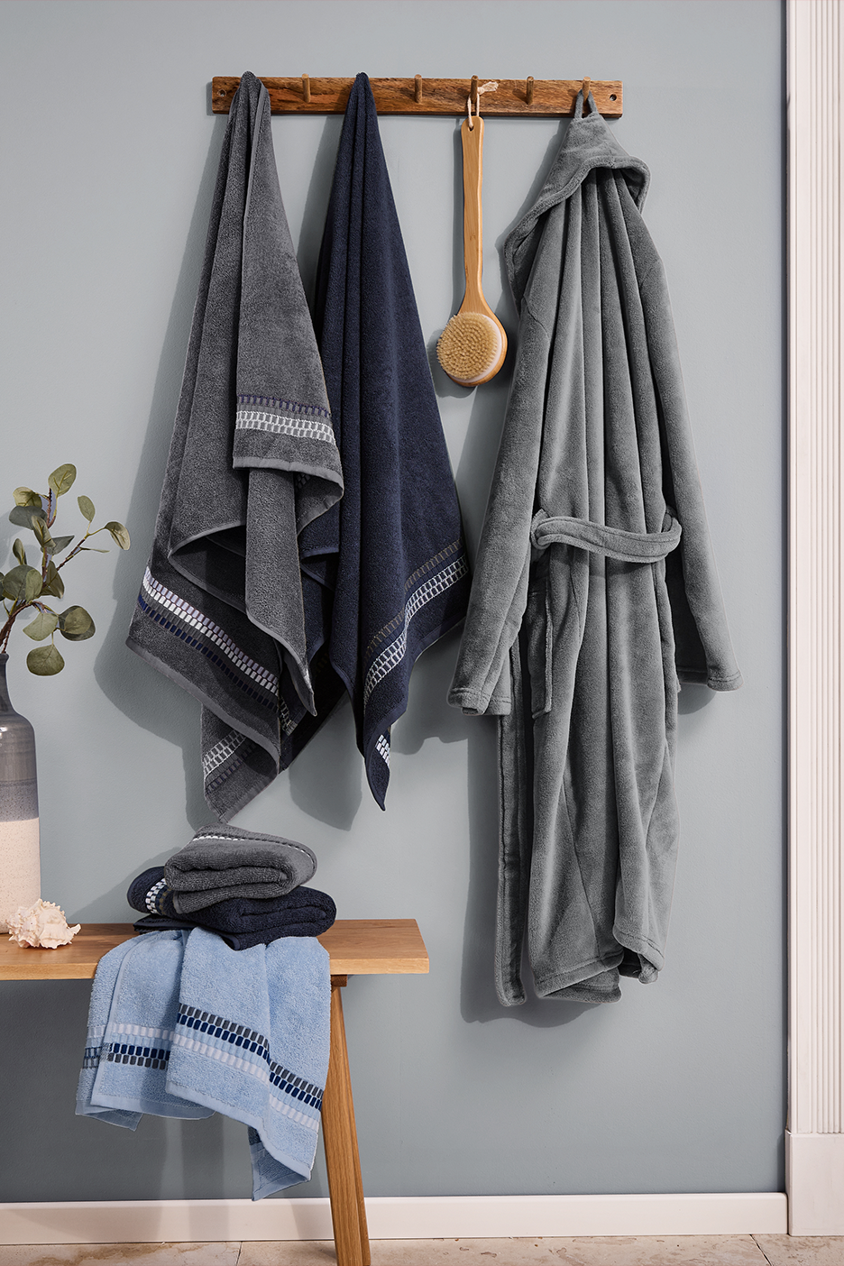 Pilki ir mėlyni rankšluosčiai bei chalatas kabo ant medinio kabliuko vonios kambaryje.