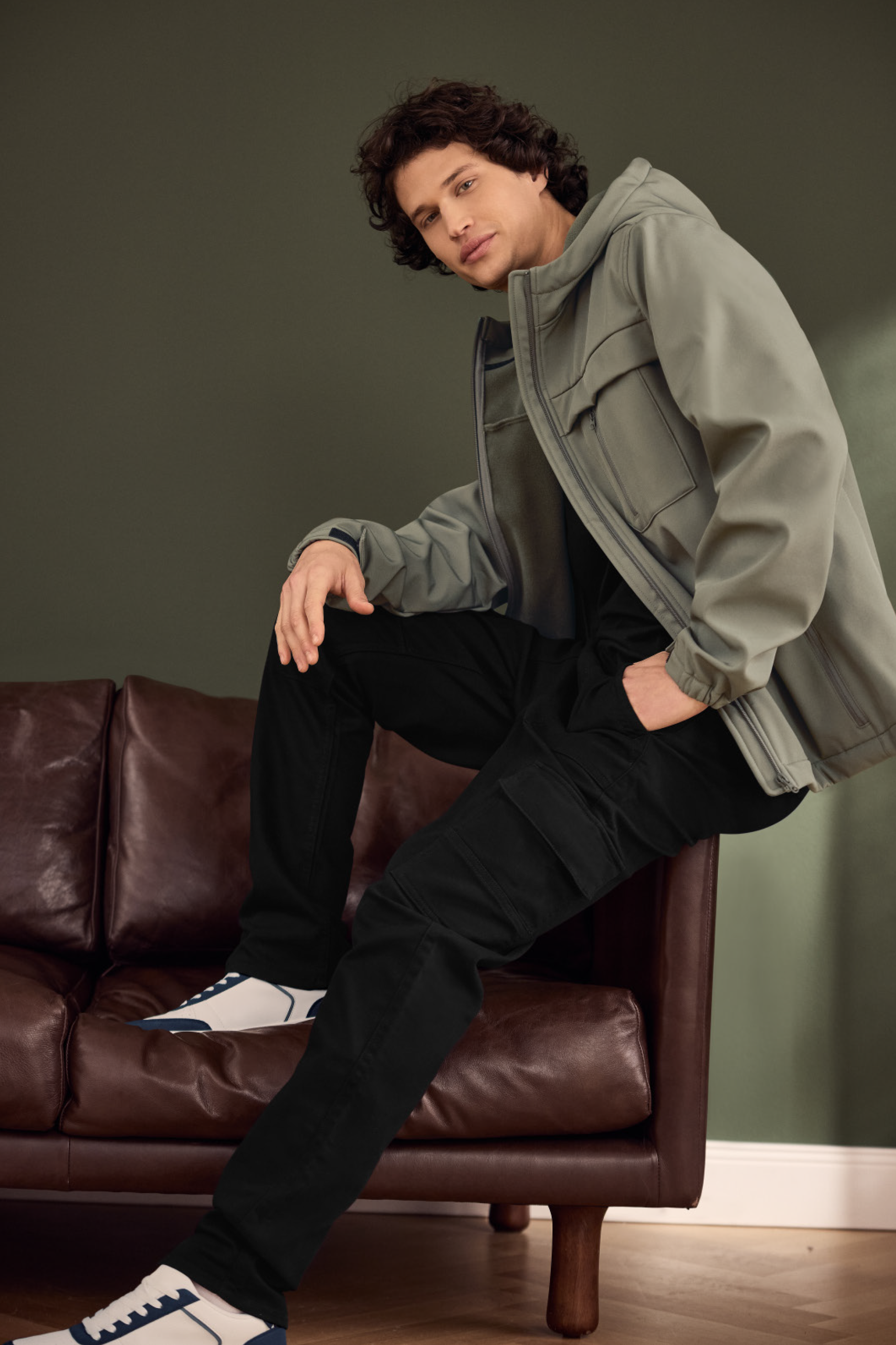 Vyras su žalia striuke, juodomis kelnėmis ir baltais sportbačiais sėdi ant rudos odinės sofos.