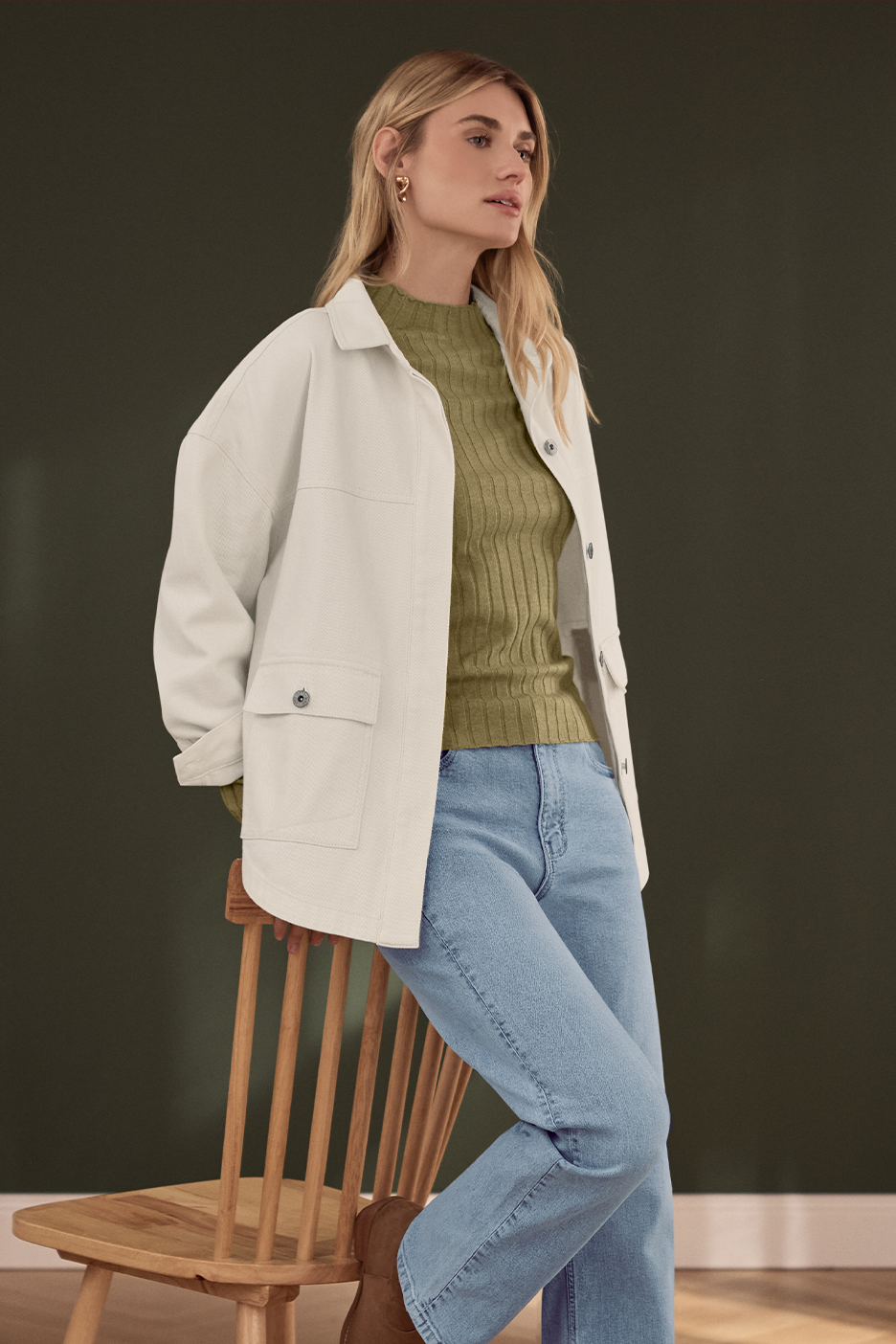 Moteris su šviesiai žaliu megztiniu, šviesiu švarkeliu ir džinsais, sėdinti ant medinės kėdės.