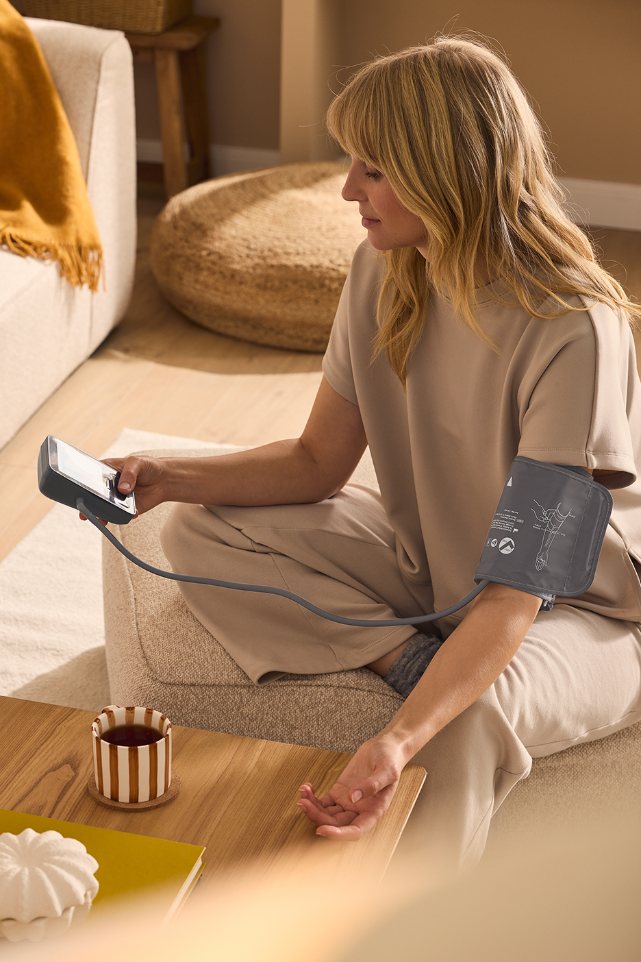 Moteris matuojasi kraujospūdį su kraujospūdžio matuokliu, sėdėdama ant sofos su puodeliu arbatos.