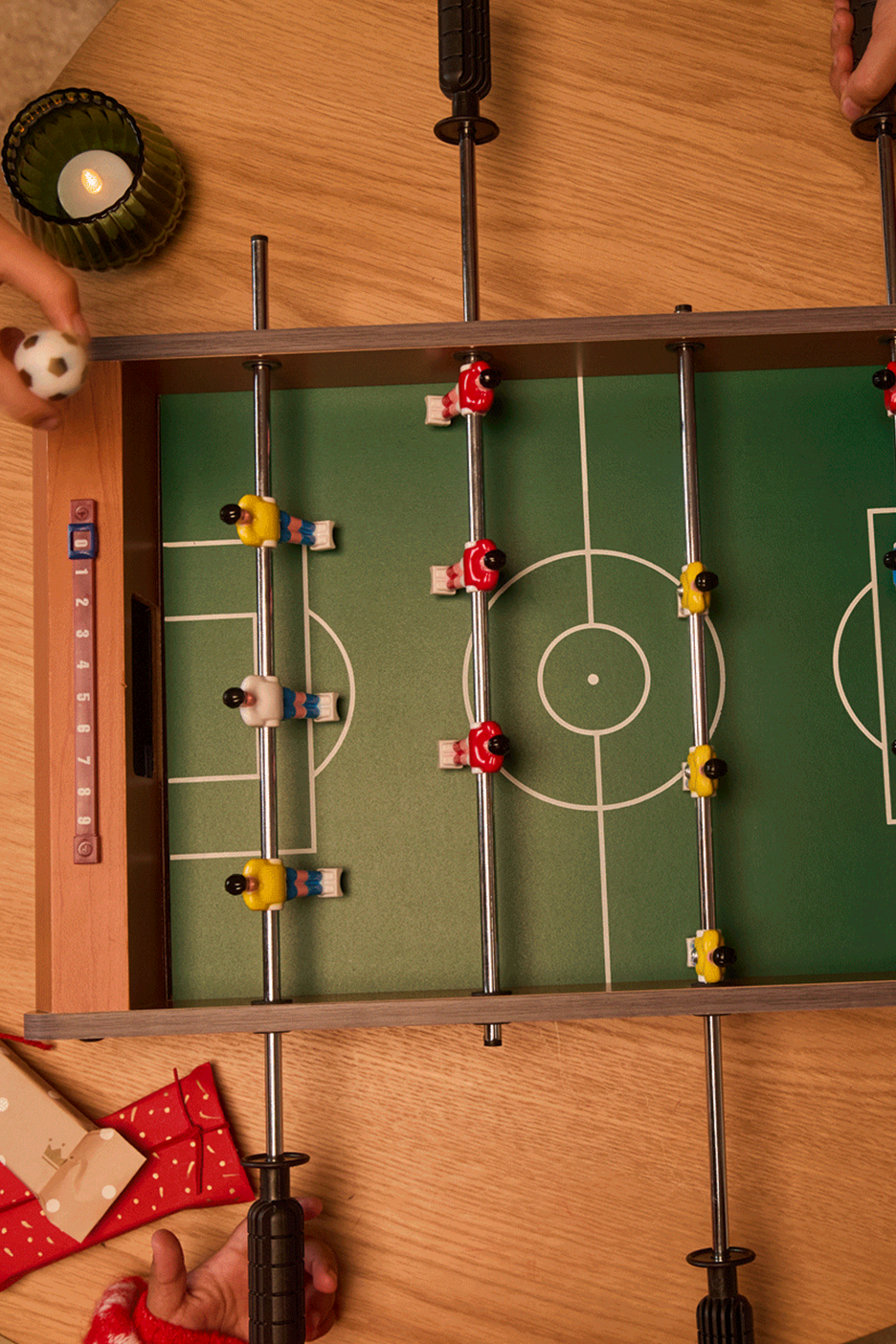 Stalo futbolas su raudonais ir geltonais žaidėjais, šalia dega žvakė ir dovanos.