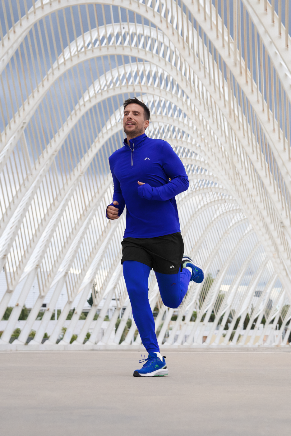 Vyras bėgioja su mėlynais marškinėliais ilgomis rankovėmis, juodais šortais ir mėlynomis tamprėmis.