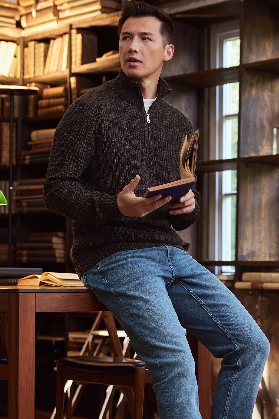 Vyras su tamsiai rudu megztiniu ir džinsais skaito knygą bibliotekoje.