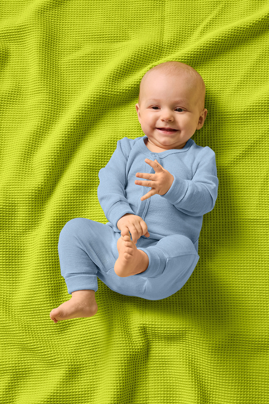 Kūdikis mėlynu kombinezonu guli ant žalios vaflinės antklodės.