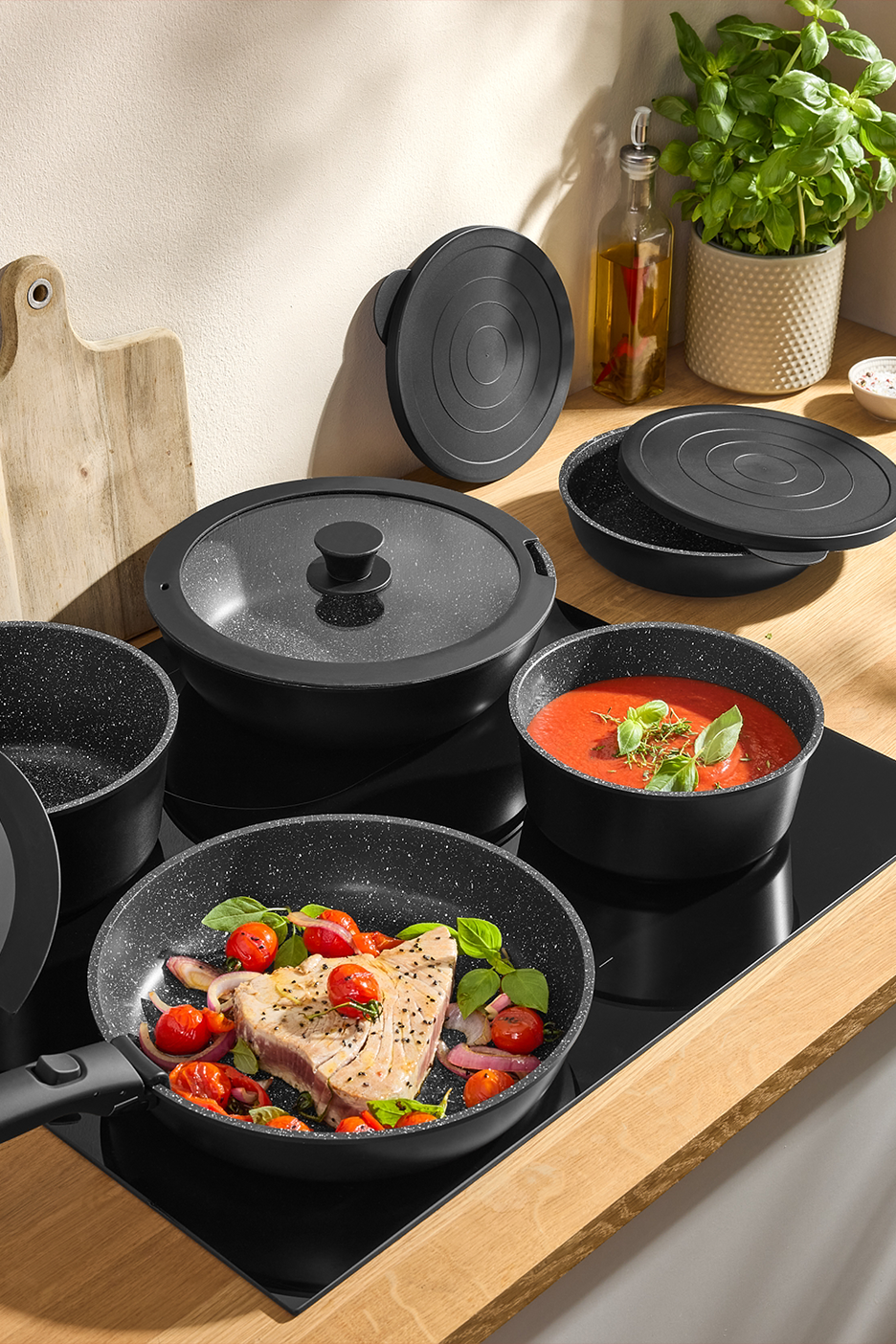 Juodų puodų ir keptuvių rinkinys su stikliniais dangčiais, ruošiamu tunu ir pomidorų sriuba.