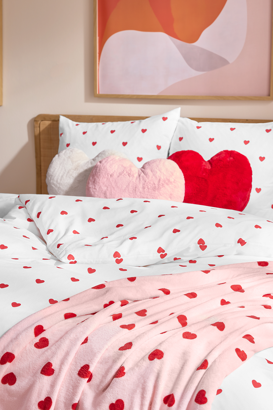 Lova su širdelėmis marginta patalyne, rožiniu pledu ir širdies formos pagalvėlėmis.