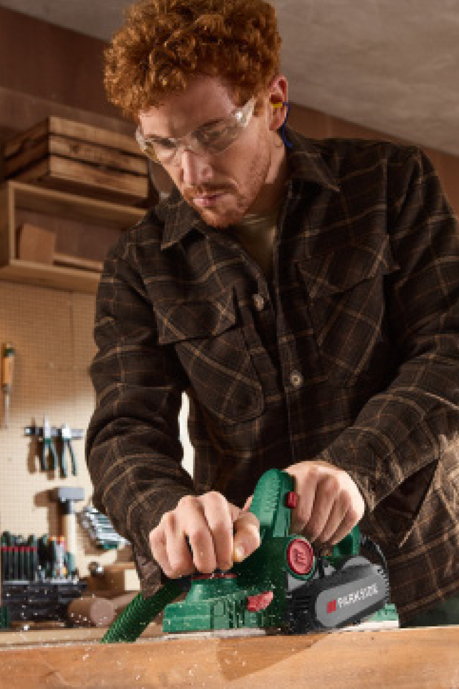 Vyras su apsauginiais akiniais ir ausų kištukais obliuoja medieną elektriniu obliumi dirbtuvėse.