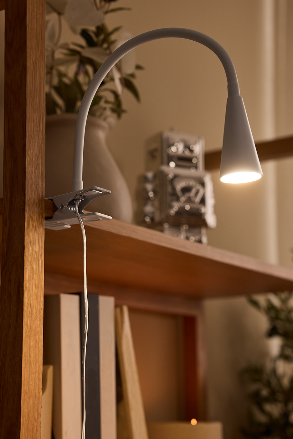 Balta prisegama lempa su lanksčiu kakleliu, apšviečianti medinę lentyną su dekoracijomis.