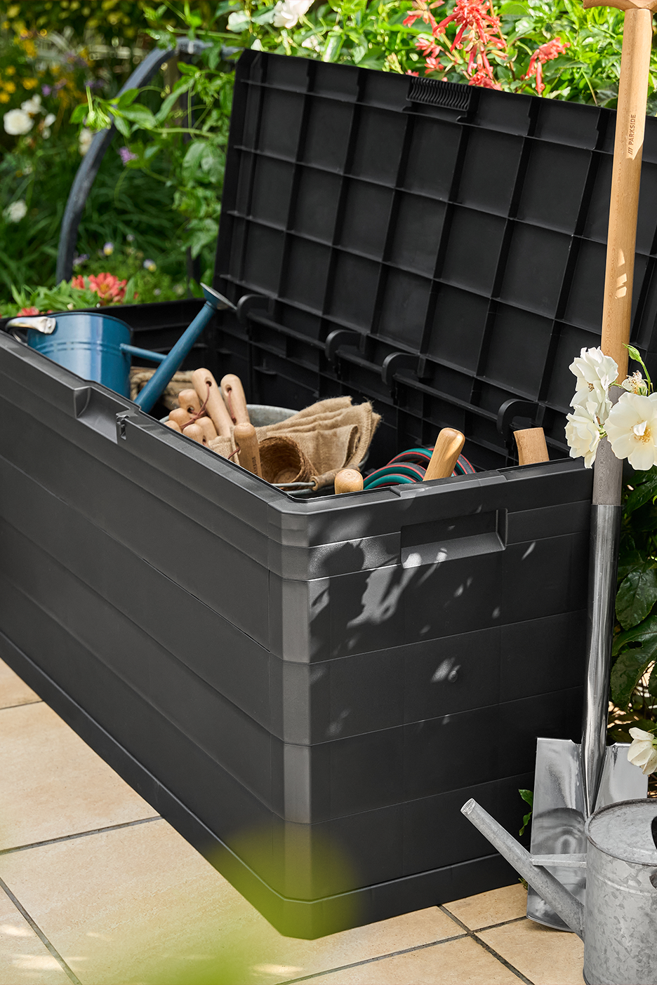 Juoda sodo daiktadėžė su atidarytu dangčiu, pilna sodo įrankių, laistytuvu ir kastuvu šalia.