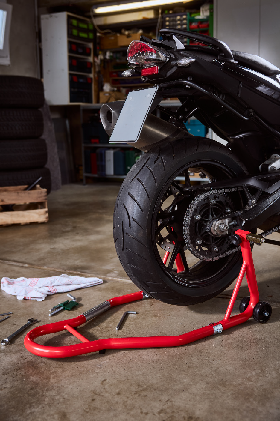Motociklas ant raudono stovo garaže, su įrankiais ir padangomis fone.