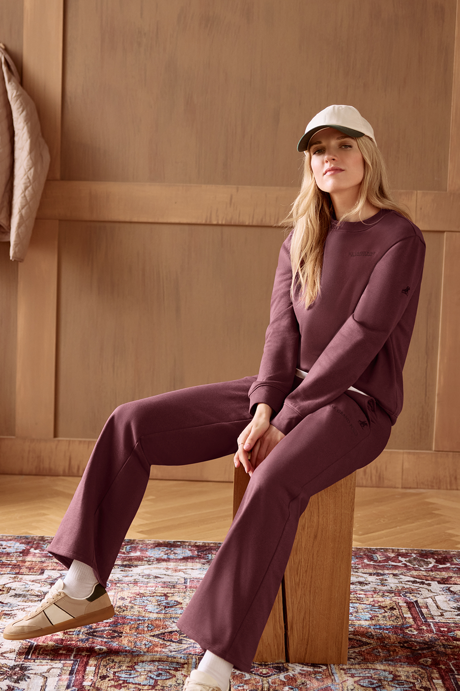 Moteris su bordo spalvos sportiniu kostiumu ir kepure sėdi ant medinio suoliuko.