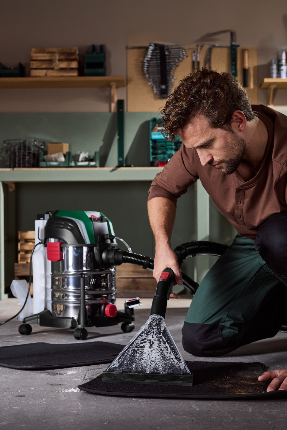 Vyras valo kilimėlį su šlapio ir sauso valymo dulkių siurbliu dirbtuvėse.