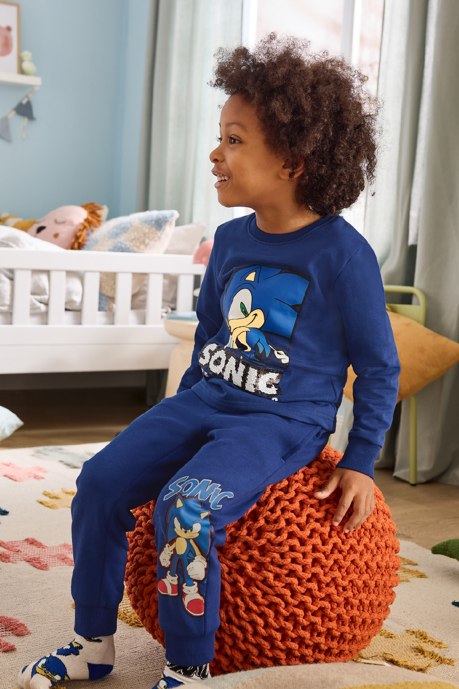 Berniukas su mėlynu sportiniu kostiumu su ežio atvaizdu sėdi ant oranžinio pufiko.