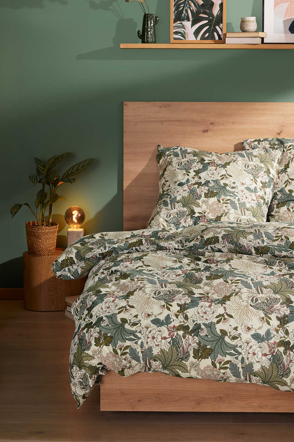 Miegamojo interjeras su medine lova, gėlėta patalyne, augalu ir šviestuvu.