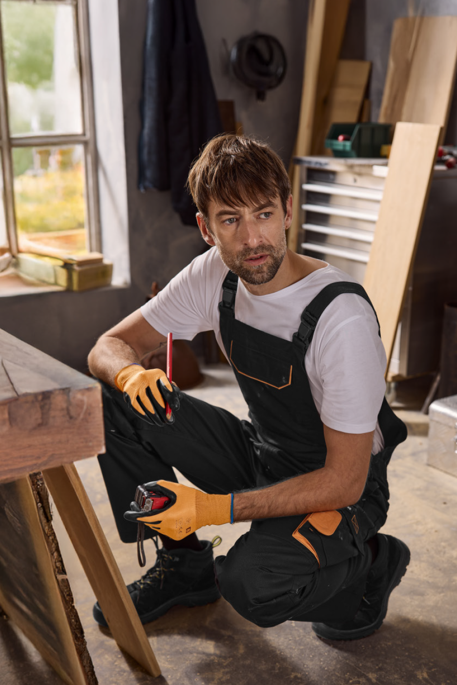 Vyras su darbo kombinezonu ir pirštinėmis dirbtuvėse.