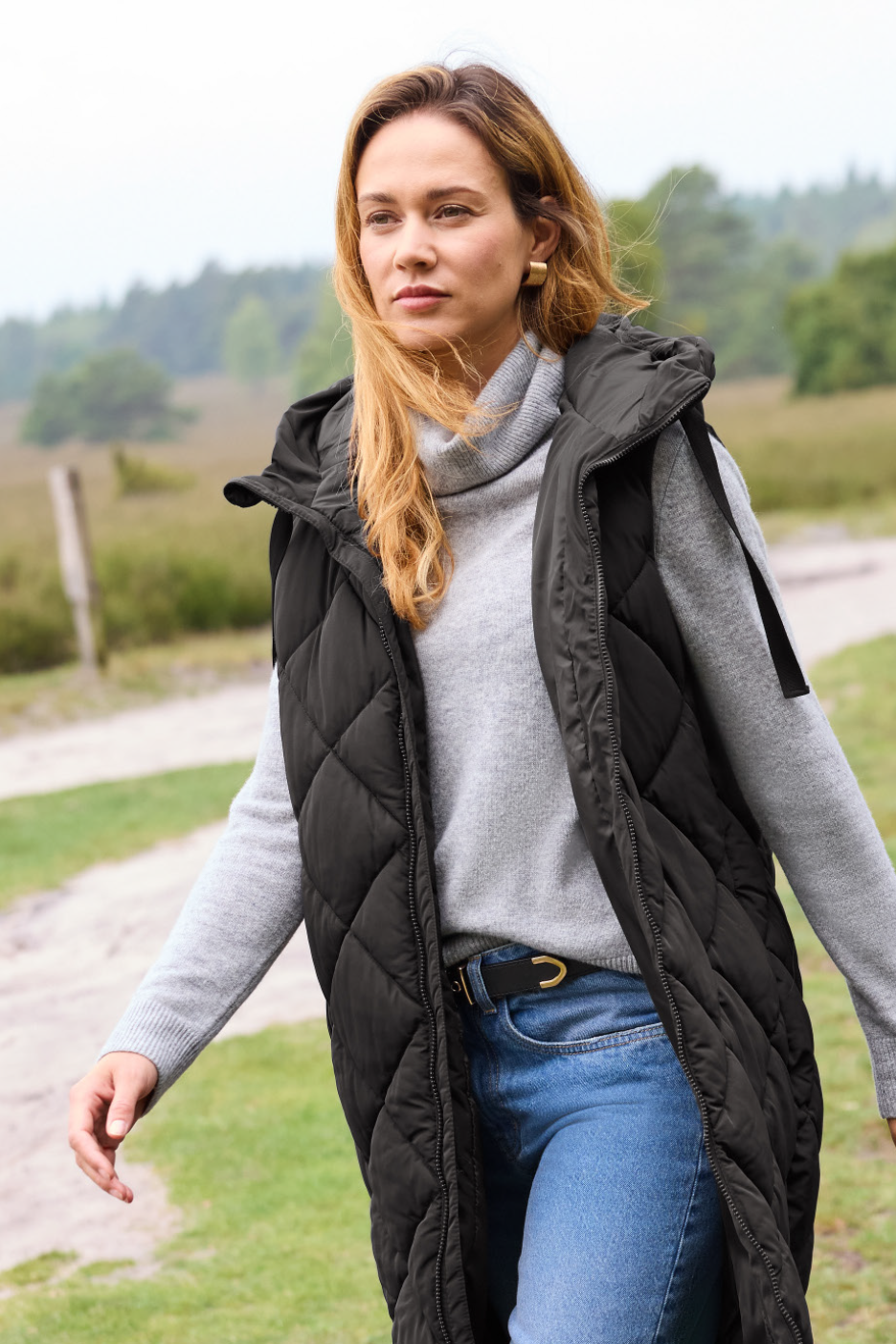 Moteris su juoda dygsniuota liemene, pilku megztiniu ir džinsais eina lauke.