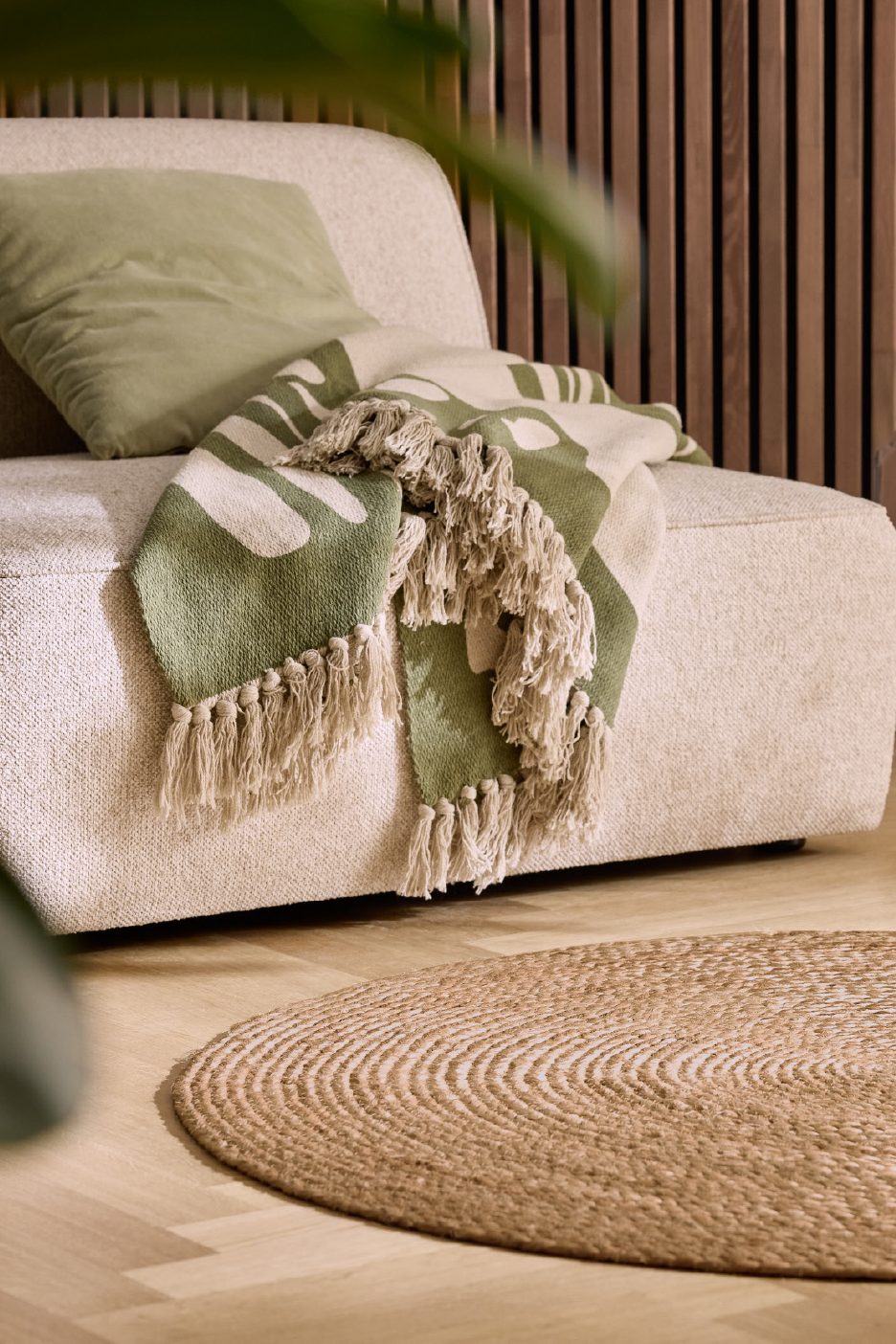 Šviesus fotelis su žaliu pagalvėle ir pledas su kutais, ant medinių grindų – apvalus kilimas.