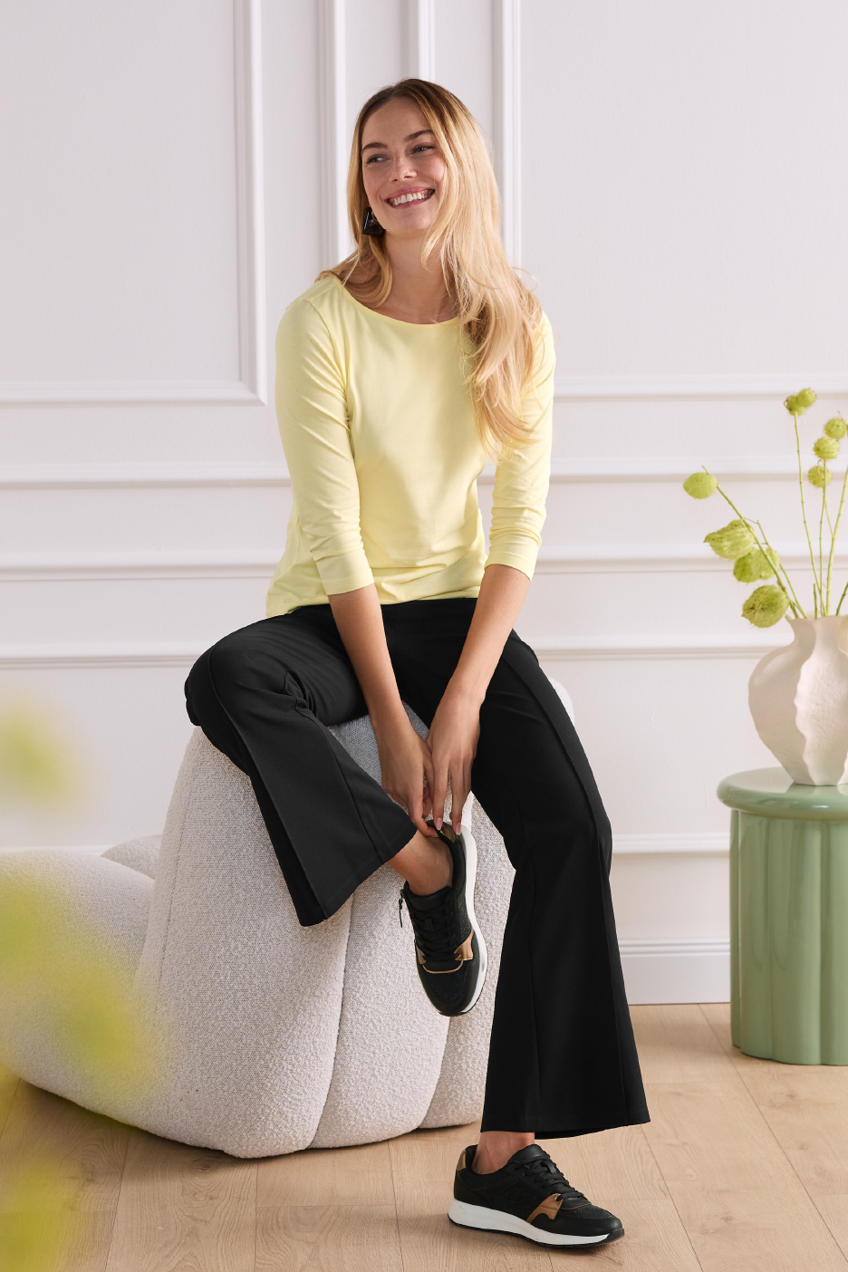 Moteris su geltonais marškinėliais ilgomis rankovėmis ir juodomis kelnėmis sėdi ant pufų.