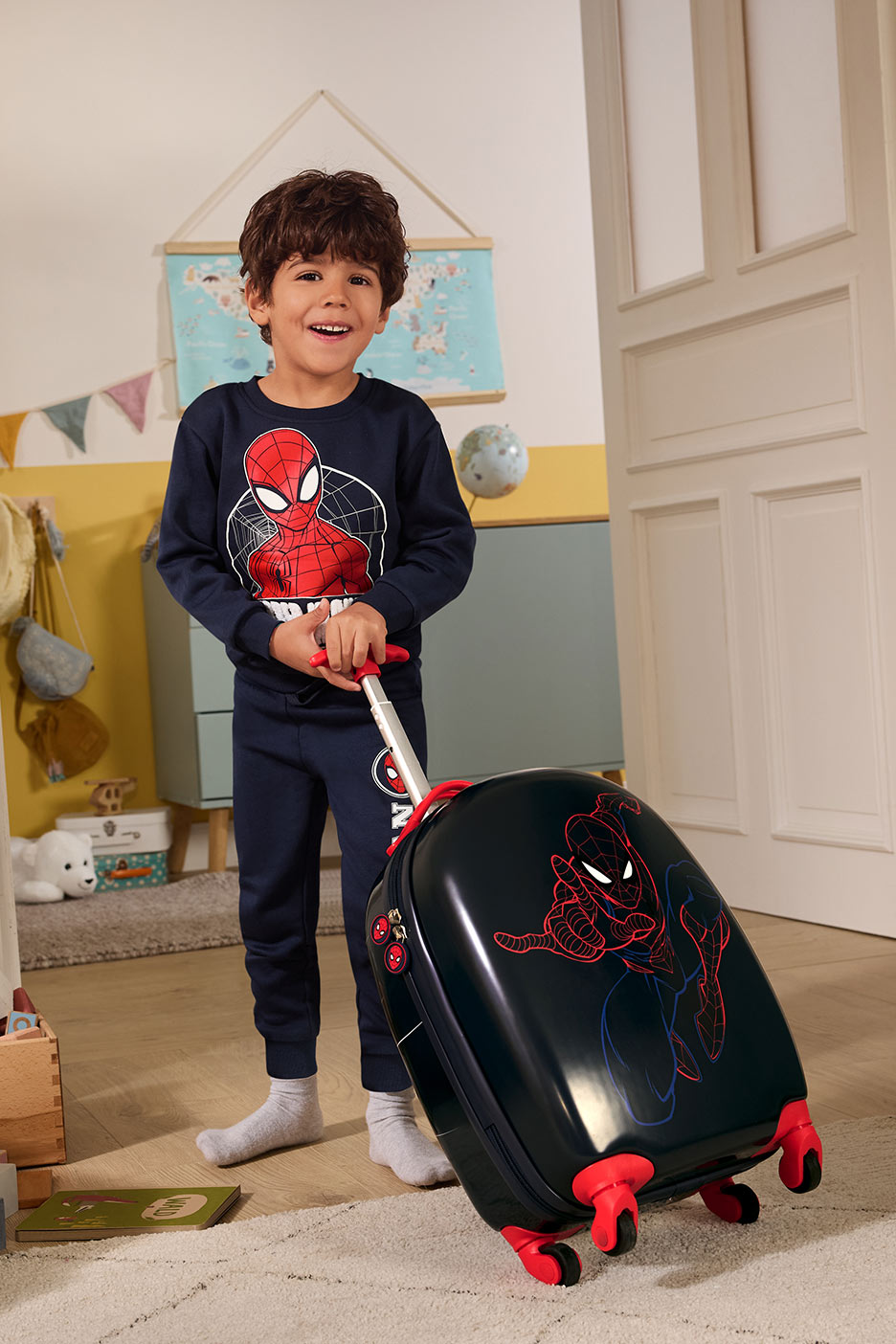 Berniukas su mėlynu megztiniu ir kelnėmis, laiko juodą lagaminą su voro herojaus atvaizdu.