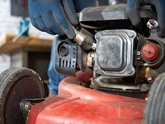 Pirštinėtos rankos keičia žvakę raudoname vejapjovyje.