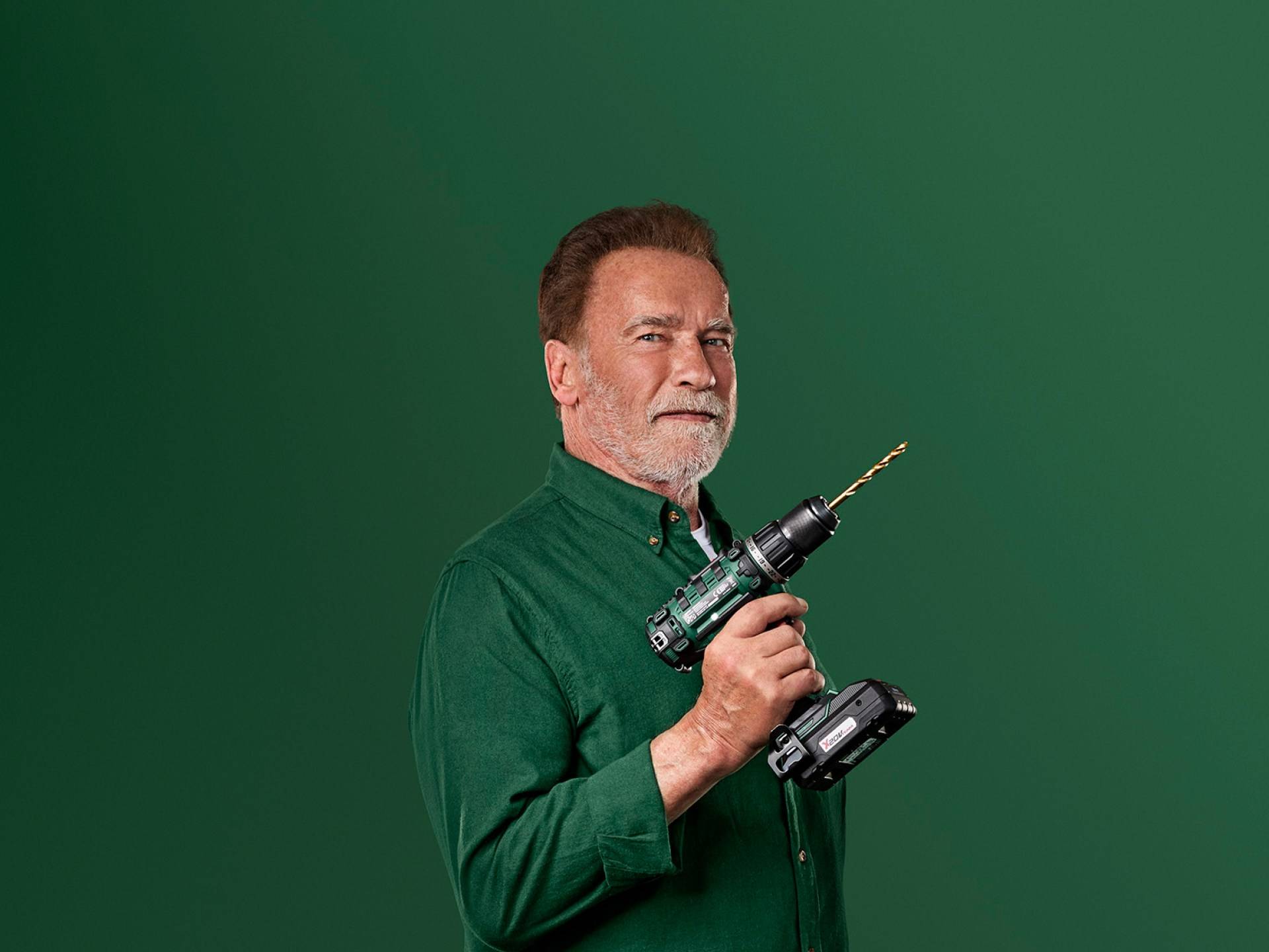 Arnoldas Schwarzeneggeris laiko žalią elektrinį grąžtą ant žalio fono.